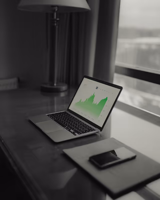 Laptop showing a green line graph on a wooden desk at AlisAlpha, alongside a phone and notebook, reflecting a modern, data-driven workflow.